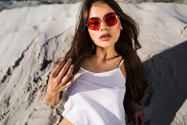 Seductora joven en gafas de sol rojas se encuentra en la arena blanca