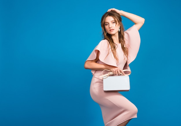 Seductora mujer sexy con estilo joven en vestido rosa de lujo, tendencia de moda de verano, estilo chic, fondo de estudio azul, sosteniendo bolso de moda