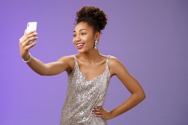 Segura, alegre, atractiva, elegante, milenaria mujer afroamericana en vestido plateado brillante tomando selfie posando lindo descarado sostener la cintura de la mano extender el brazo grabar mensaje de video durante la noche.