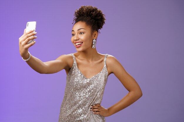Segura, alegre, atractiva, elegante, milenaria mujer afroamericana en vestido plateado brillante tomando selfie posando lindo descarado sostener la cintura de la mano extender el brazo grabar mensaje de video durante la noche.