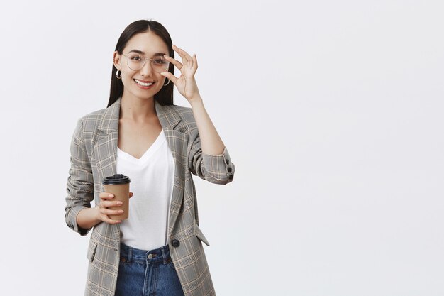 Segura y alegre mujer guapa con gafas y elegante chaqueta, tocando el borde y sonriendo ampliamente mientras sostiene una taza de café, bebiendo bebidas