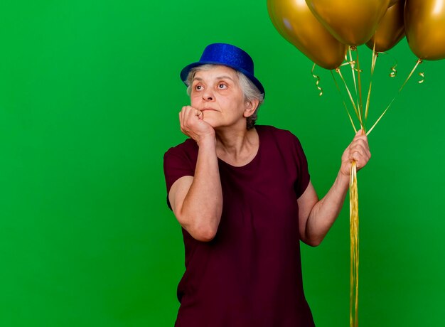 Segura anciana con gorro de fiesta sostiene globos de helio y pone la mano en la barbilla mirando al costado en verde