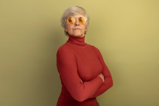 Segura anciana vestida con un suéter de cuello alto rojo y gafas de sol de pie con una postura cerrada en la vista de perfil aislada en la pared verde oliva con espacio de copia