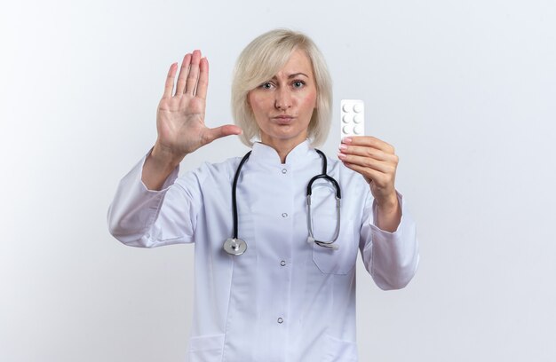 Segura doctora eslava adulta en bata médica con estetoscopio gesticulando señal de stop y sosteniendo la tableta de medicina en blister aislado sobre fondo blanco con espacio de copia