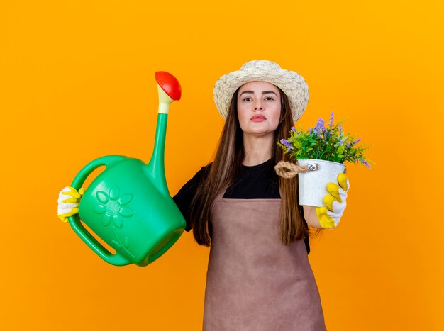 Segura hermosa chica jardinero con uniforme y sombrero de jardinería con guantes levantando regadera y sosteniendo flores en flwerpot en cámara aislada sobre fondo naranja