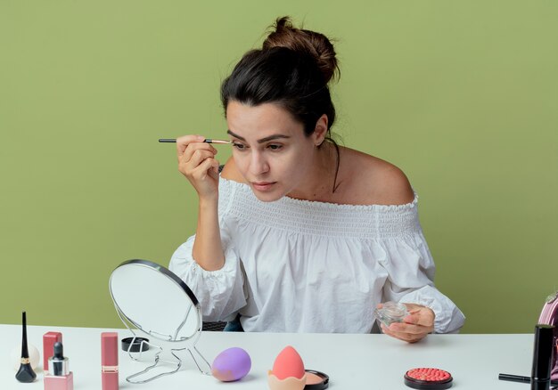 Segura hermosa chica se sienta a la mesa con herramientas de maquillaje mira espejo aplicando sombra de ojos con pincel de maquillaje aislado en la pared verde