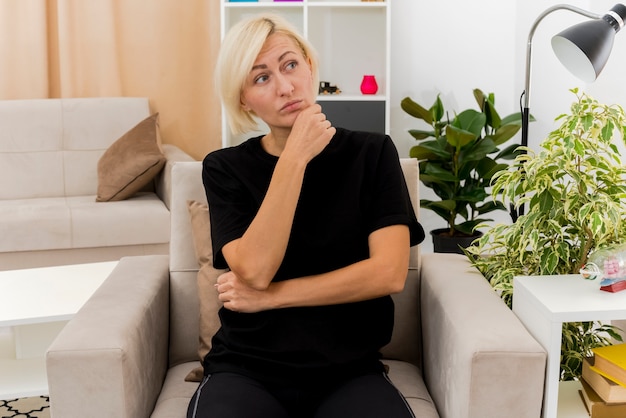 Foto gratuita segura hermosa mujer rusa rubia sentada en un sillón poniendo la mano en la barbilla mirando de lado dentro de la sala de estar