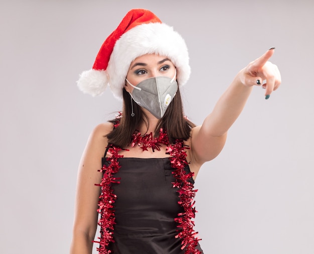 Segura joven bastante caucásica vestida con gorro de Papá Noel y máscara protectora guirnalda de oropel alrededor del cuello mirando y apuntando al lado aislado sobre fondo blanco.