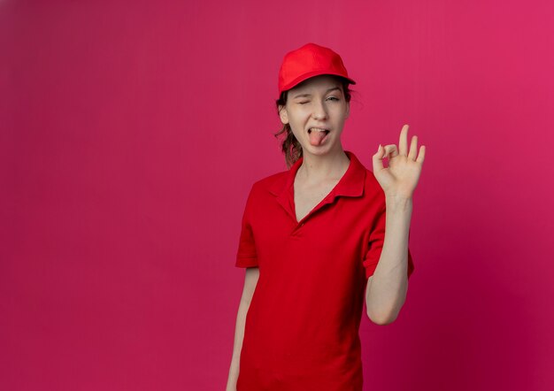 Segura joven bonita repartidora con uniforme rojo y gorra haciendo el signo de ok guiñando un ojo y mostrando la lengua aislada sobre fondo carmesí con espacio de copia