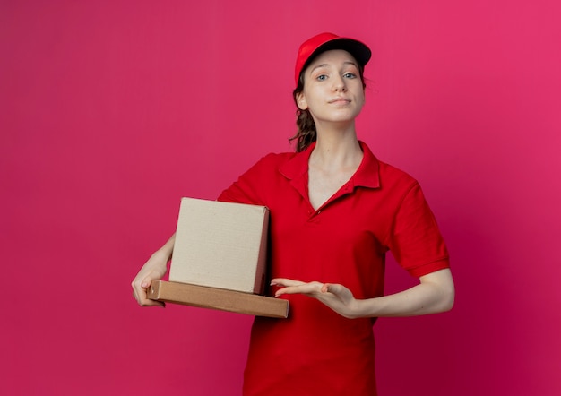 Segura joven bonita repartidora con uniforme rojo y gorra sosteniendo y apuntando con la mano a la caja de cartón y el paquete de pizza aislado sobre fondo carmesí con espacio de copia