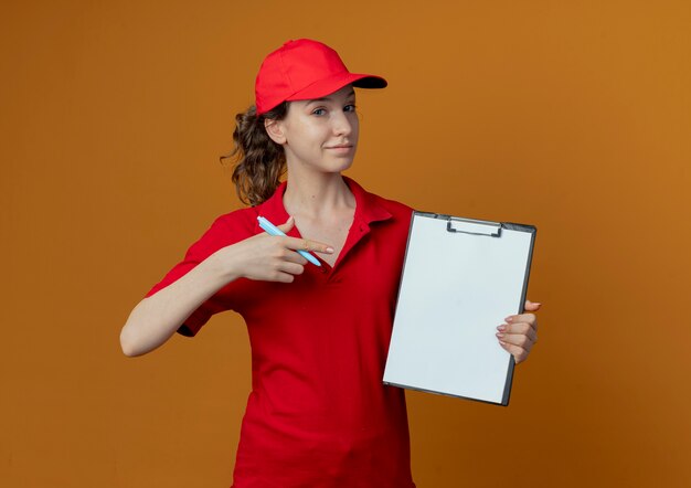 Segura joven bonita repartidora en uniforme rojo y gorra sosteniendo bolígrafo y portapapeles y apuntando al portapapeles aislado sobre fondo naranja con espacio de copia