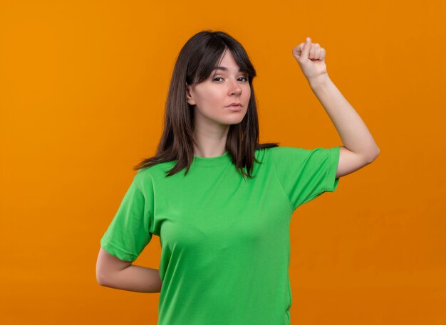 Segura joven caucásica en camisa verde levanta el puño y mira a la cámara sobre fondo naranja aislado