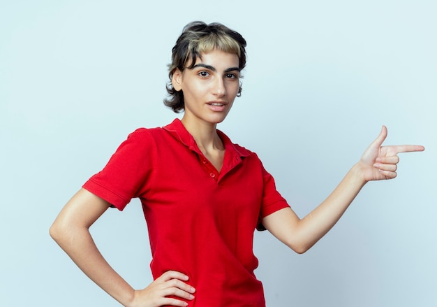 Foto gratuita segura joven caucásica con corte de pelo pixie apuntando al lado con otra mano en la cintura aislada sobre fondo blanco