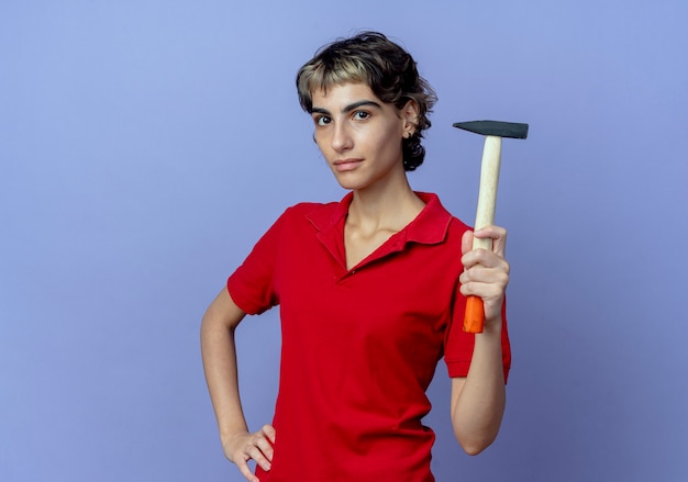 Segura joven caucásica con corte de pelo pixie sosteniendo un martillo y poniendo la mano en la cintura aislada sobre fondo púrpura con espacio de copia