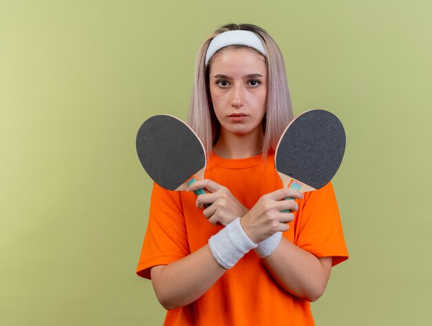 Segura joven caucásica deportiva con tirantes con diadema y muñequeras sostiene raquetas de ping-pong
