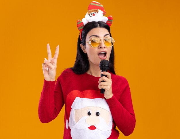 Segura joven caucásica vistiendo santa claus diadema y suéter con gafas hablando por micrófono mirando a cámara haciendo el signo de la paz aislado sobre fondo naranja