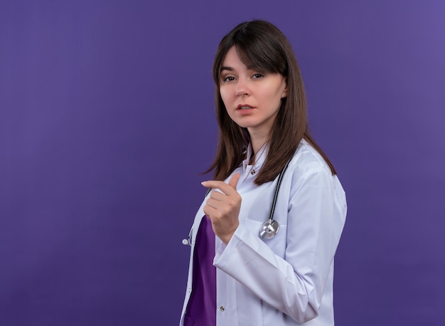 Segura joven doctora en bata médica con estetoscopio se encuentra de lado y mira a la cámara sobre fondo violeta aislado con espacio de copia