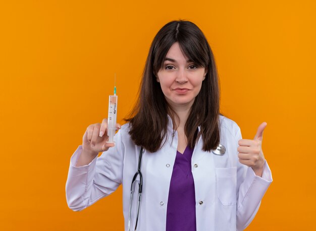 Segura joven doctora en bata médica con estetoscopio sostiene jeringa y pulgar hacia arriba sobre fondo naranja aislado con espacio de copia