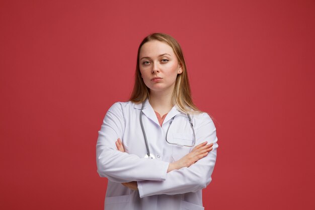 Segura joven doctora rubia vistiendo bata médica y estetoscopio alrededor del cuello de pie con postura cerrada