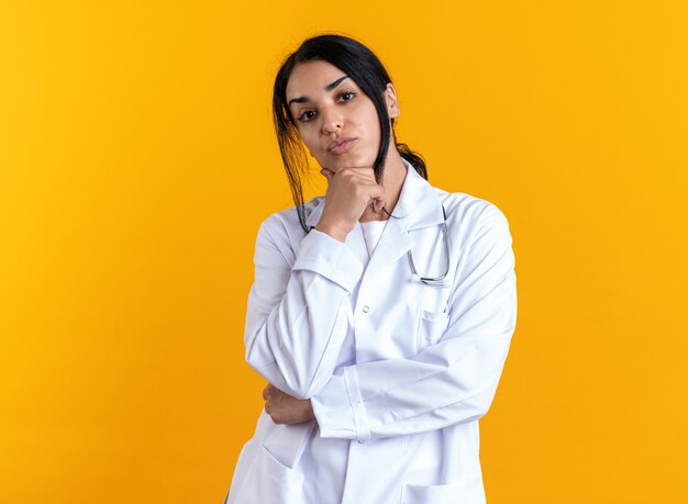 Segura joven doctora vistiendo bata médica con estetoscopio agarró la barbilla aislada en la pared amarilla