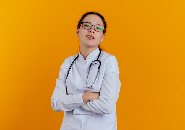 Segura joven doctora vistiendo bata médica y estetoscopio con gafas cruzando las manos aisladas