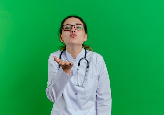 Segura joven doctora vistiendo bata médica y estetoscopio y gafas enviando beso golpe manteniendo la mano en el aire aislada en la pared verde con espacio de copia