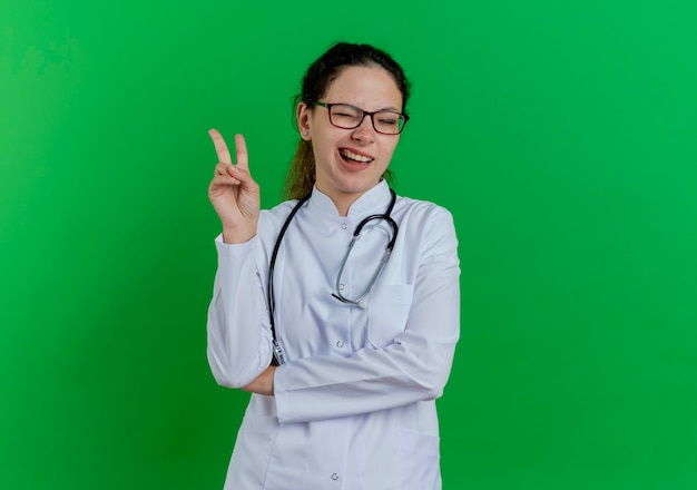 Segura joven doctora vistiendo bata médica y estetoscopio y gafas haciendo el signo de la paz guiñando un ojo aislado en la pared verde con espacio de copia