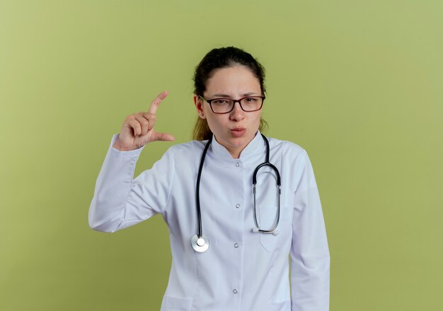 Segura joven doctora vistiendo bata médica y un estetoscopio con gafas que muestran el tamaño aislado en la pared verde oliva