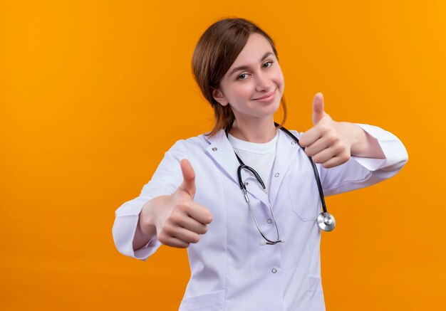 Segura joven doctora vistiendo bata médica y un estetoscopio mostrando los pulgares hacia arriba en el espacio naranja aislado con espacio de copia