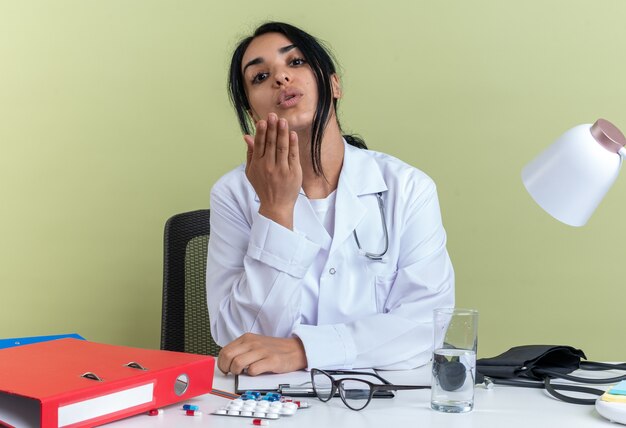 Segura joven doctora vistiendo bata médica con estetoscopio se sienta en el escritorio con herramientas médicas que muestran gesto de beso aislado en la pared verde oliva