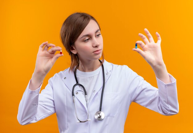 Segura joven doctora vistiendo bata médica y estetoscopio sosteniendo cápsulas médicas y mirándolas en un espacio naranja aislado