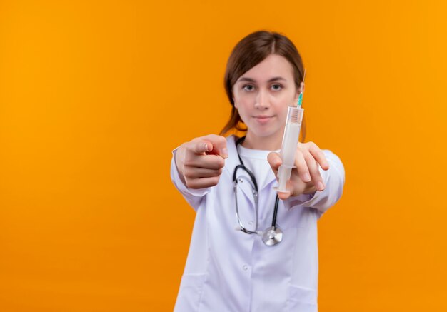 Segura joven doctora vistiendo bata médica y un estetoscopio sosteniendo una jeringa y apuntando a un espacio naranja aislado con espacio de copia