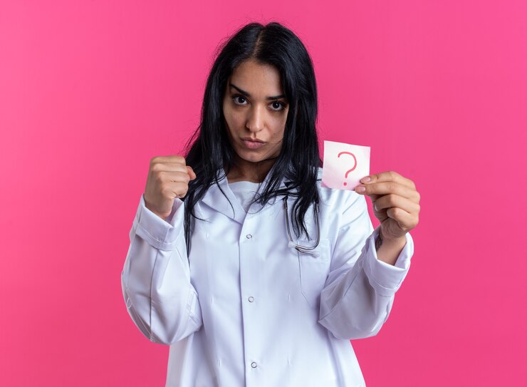 Foto gratuito segura joven doctora vistiendo bata médica con estetoscopio sosteniendo papel de nota en signo de interrogación aislado en la pared rosa