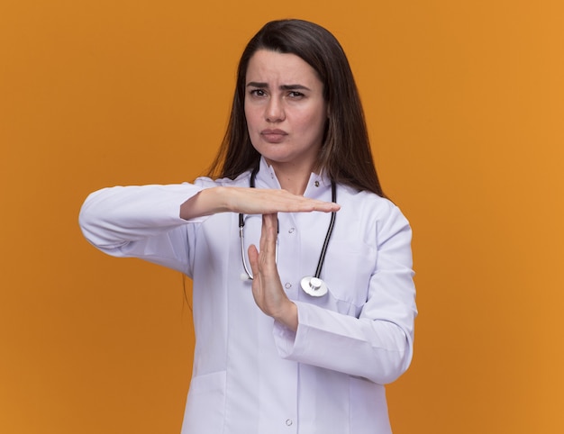 Segura joven doctora vistiendo bata médica con gestos de estetoscopio tiempo de espera firmar en naranja