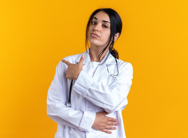 Segura joven doctora vistiendo bata médica con puntos de estetoscopio detrás aislado en la pared amarilla con espacio de copia