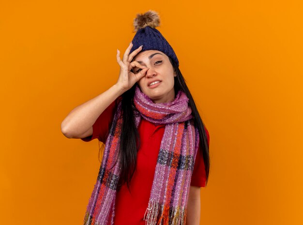 Segura joven enferma vistiendo gorro y bufanda de invierno haciendo gesto de mirada mirando al frente aislado en la pared naranja