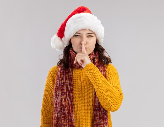 Segura joven eslava con gorro de Papá Noel y bufanda alrededor del cuello haciendo gesto de silencio aislado en la pared blanca con espacio de copia