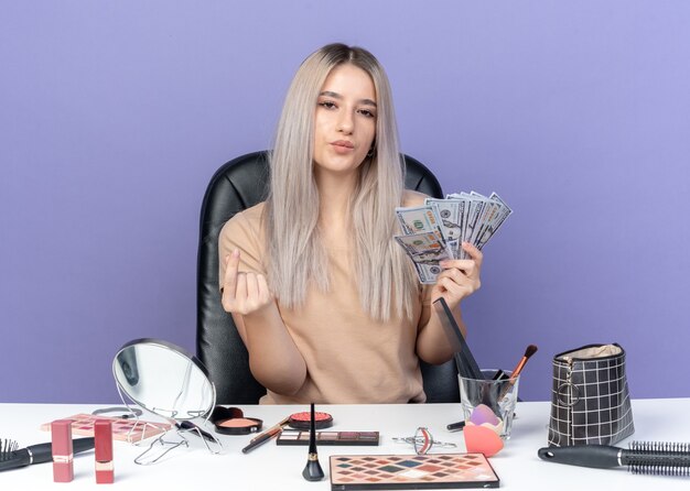 Segura joven hermosa se sienta a la mesa con herramientas de maquillaje con dinero en efectivo mostrando gesto de propina aislado en la pared azul