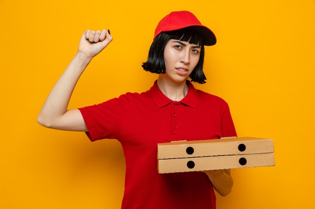Segura joven mujer caucásica de entrega sosteniendo cajas de pizza y levantando su puño