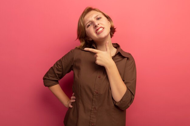 Segura joven rubia mirando manteniendo la mano en la cintura mostrando los dientes apuntando al lado aislado en la pared rosa