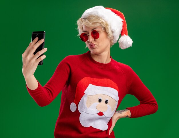 Segura joven rubia con sombrero de navidad y suéter de navidad de santa claus con gafas manteniendo la mano en la cintura tomando selfie con labios fruncidos aislados sobre fondo verde