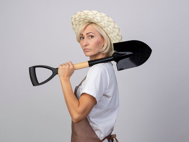 Segura de mediana edad jardinero rubio mujer en uniforme con sombrero de pie en la vista de perfil mirando al frente sosteniendo la pala en el hombro aislado en la pared blanca