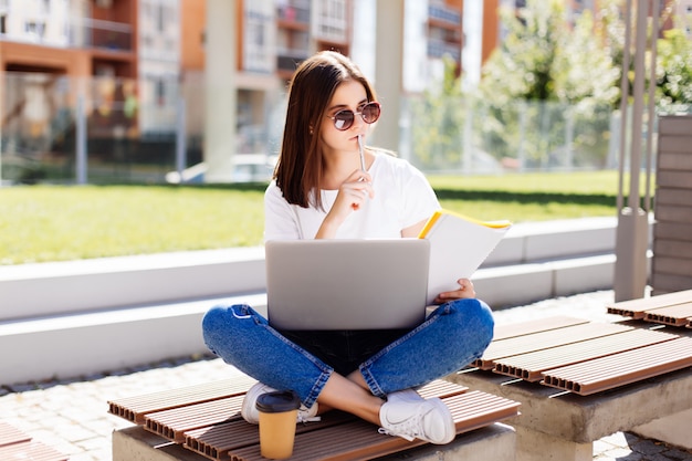 Segura mujer atractiva sentada en el banco en el parque y mensajes en las redes sociales mientras escribía en el cuaderno durante el descanso para tomar café