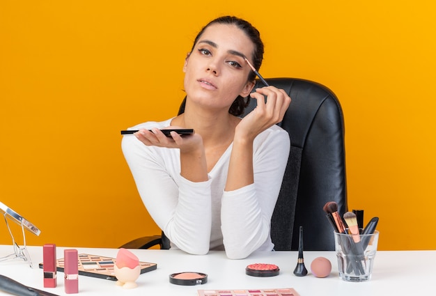 Segura de mujer bastante caucásica sentada a la mesa con herramientas de maquillaje sosteniendo la paleta de sombra de ojos y aplicando sombra de ojos con pincel de maquillaje aislado en la pared naranja con espacio de copia