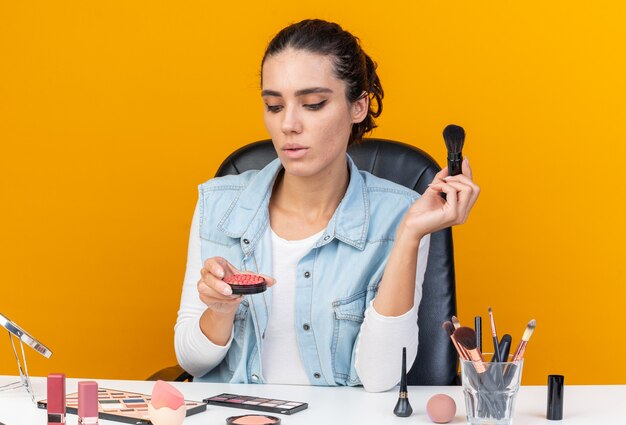 Segura de mujer bastante caucásica sentada a la mesa con herramientas de maquillaje sosteniendo pincel de maquillaje y mirando rubor aislado en pared naranja con espacio de copia