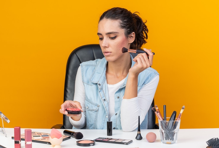 Foto gratuito segura de mujer bastante caucásica sentada a la mesa con herramientas de maquillaje sosteniendo pincel de maquillaje mirando rubor