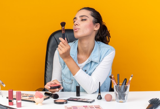 Segura de mujer bastante caucásica sentada a la mesa con herramientas de maquillaje sosteniendo rubor y mirando pincel de maquillaje