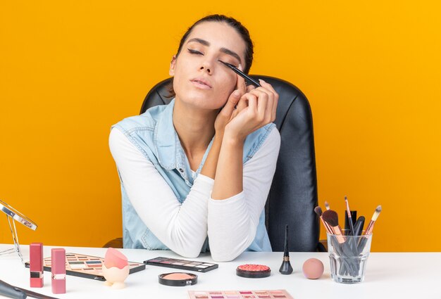 Segura de mujer bastante caucásica sentada con los ojos cerrados en la mesa con herramientas de maquillaje aplicando delineador de ojos