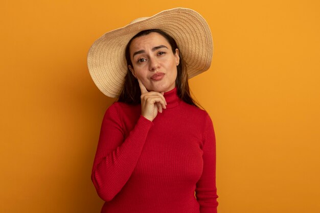 Segura de mujer bonita con sombrero de playa pone la mano en la barbilla aislada en la pared naranja