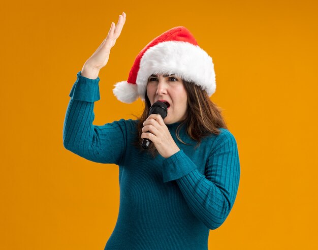 Segura mujer caucásica adulta con gorro de Papá Noel tiene micrófono fingiendo cantar aislado en la pared naranja con espacio de copia